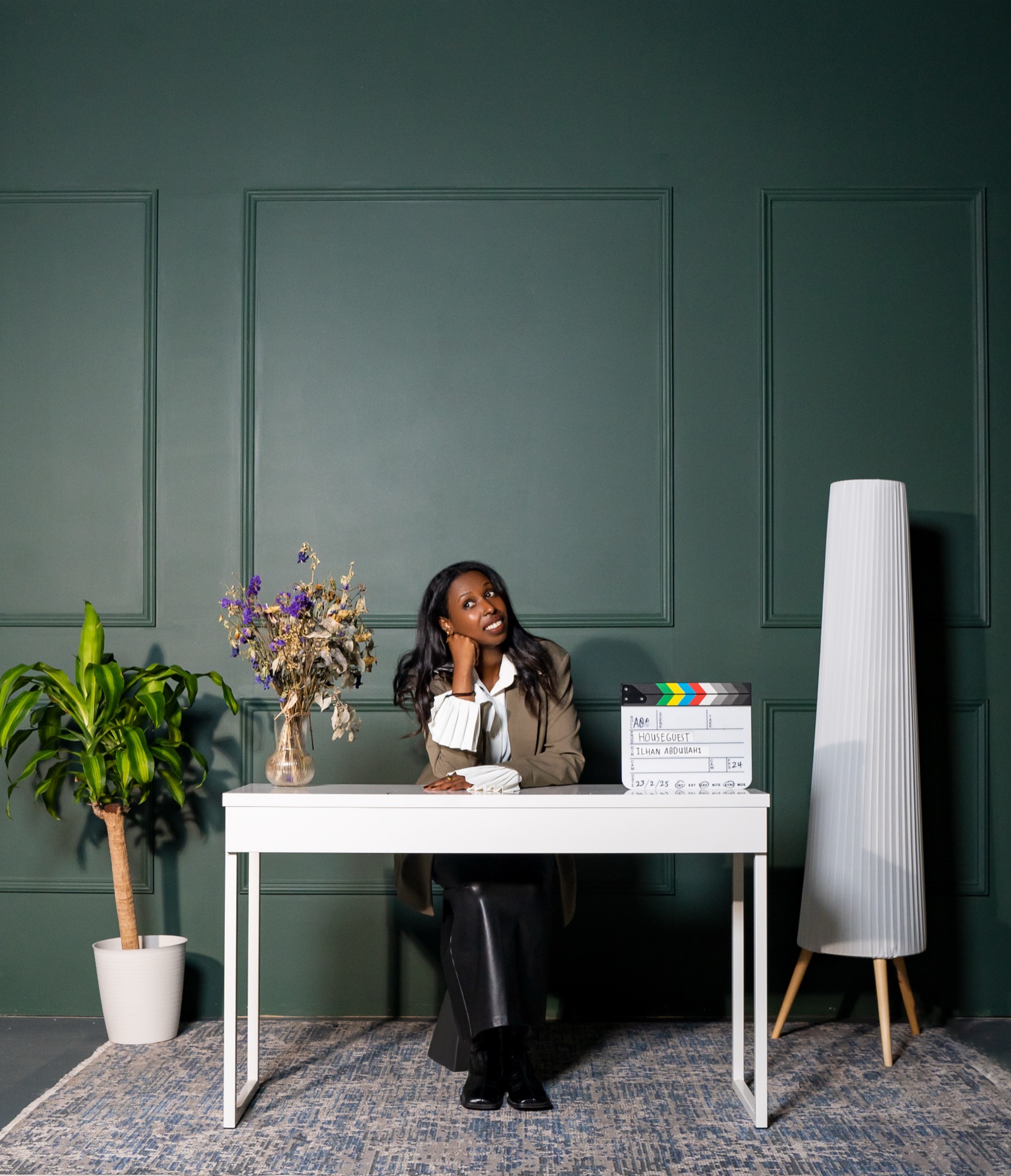 Ilhan Abdullahi seated at a white desk, smiling, beside a clapperboard marked with her name.
