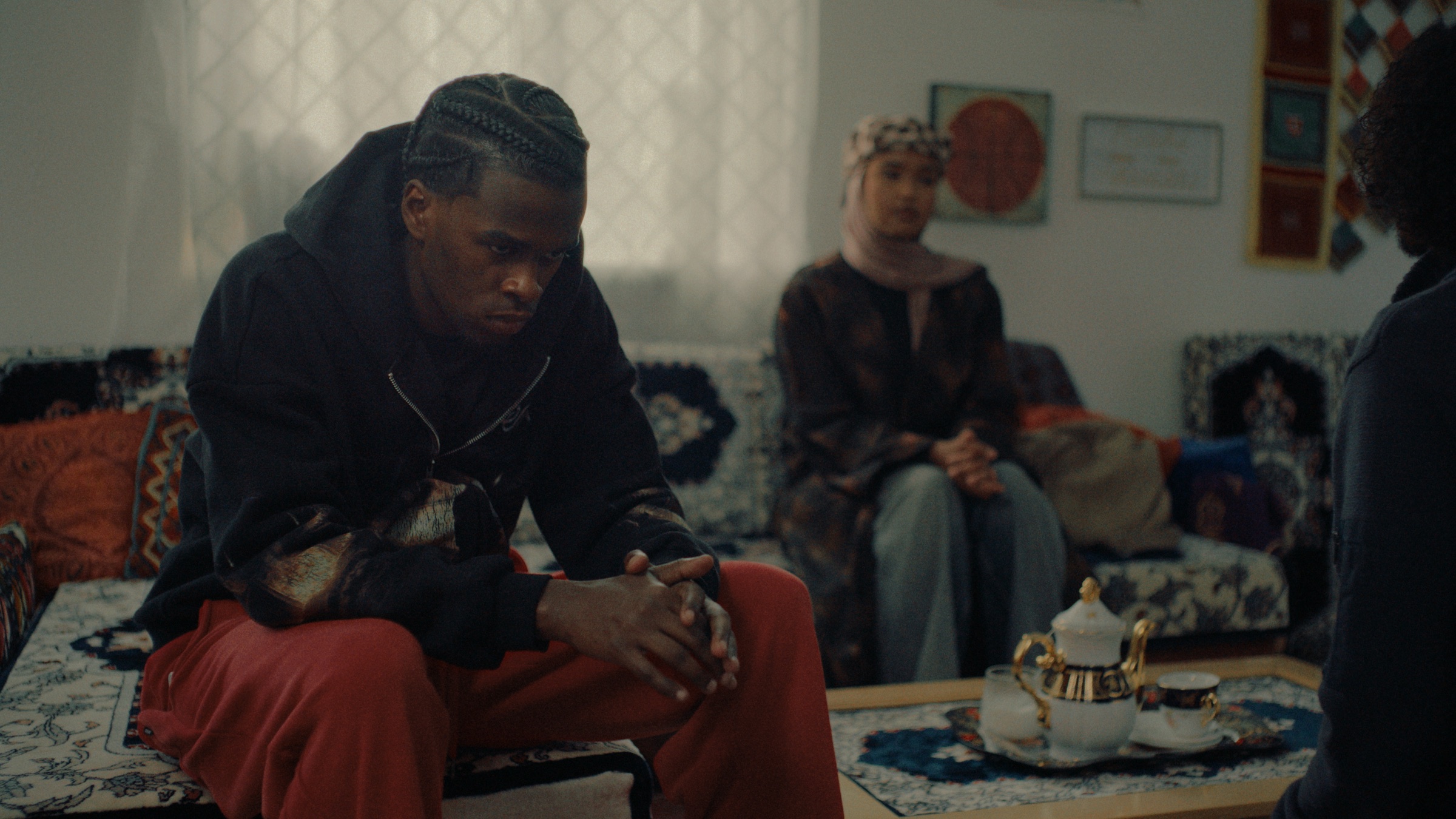Hassan sits hunched forward on a couch, hands clasped, while a woman watches from across the room.