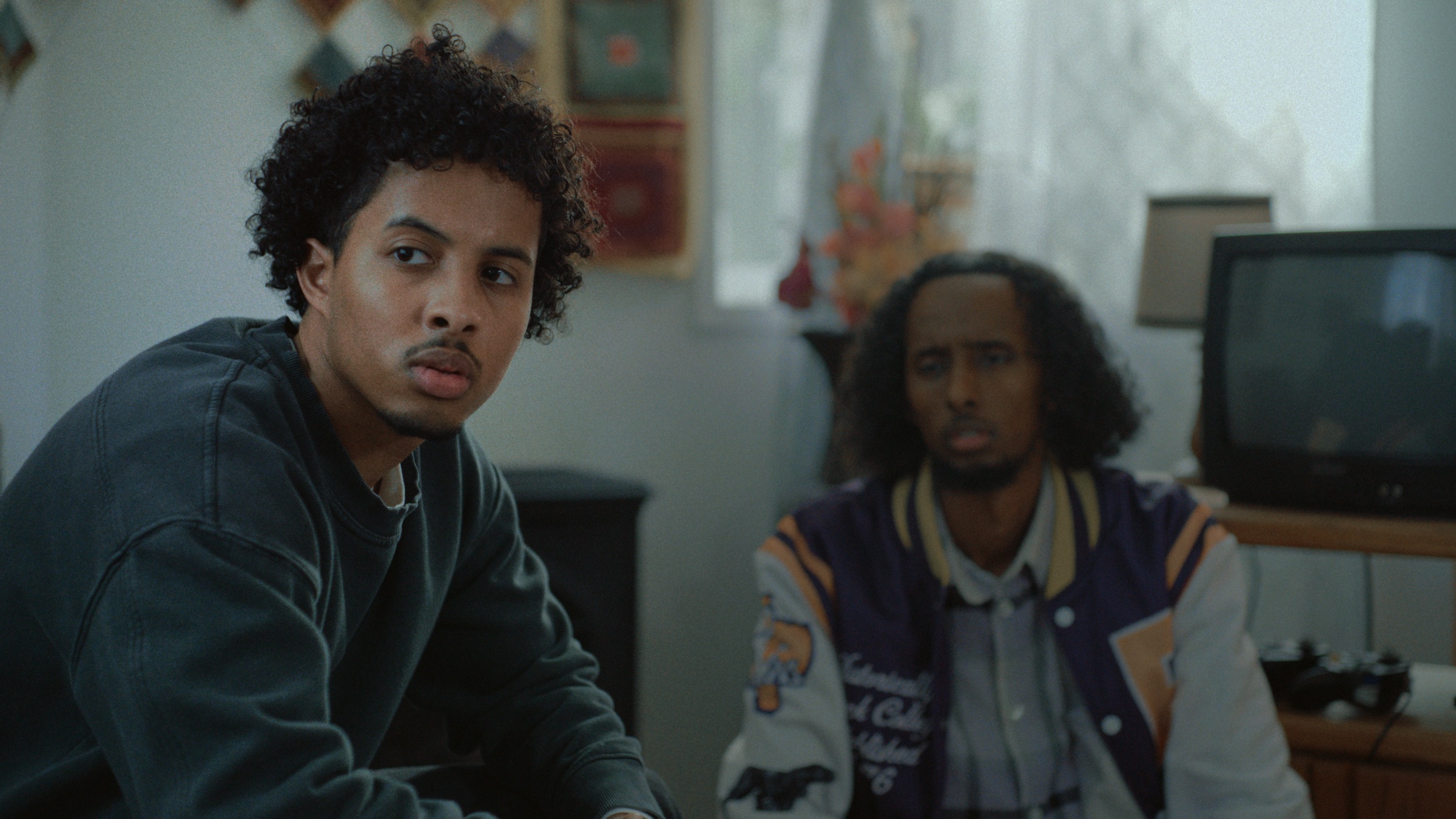 Two young men in a living room, one looking over his shoulder, the other watching from a couch.