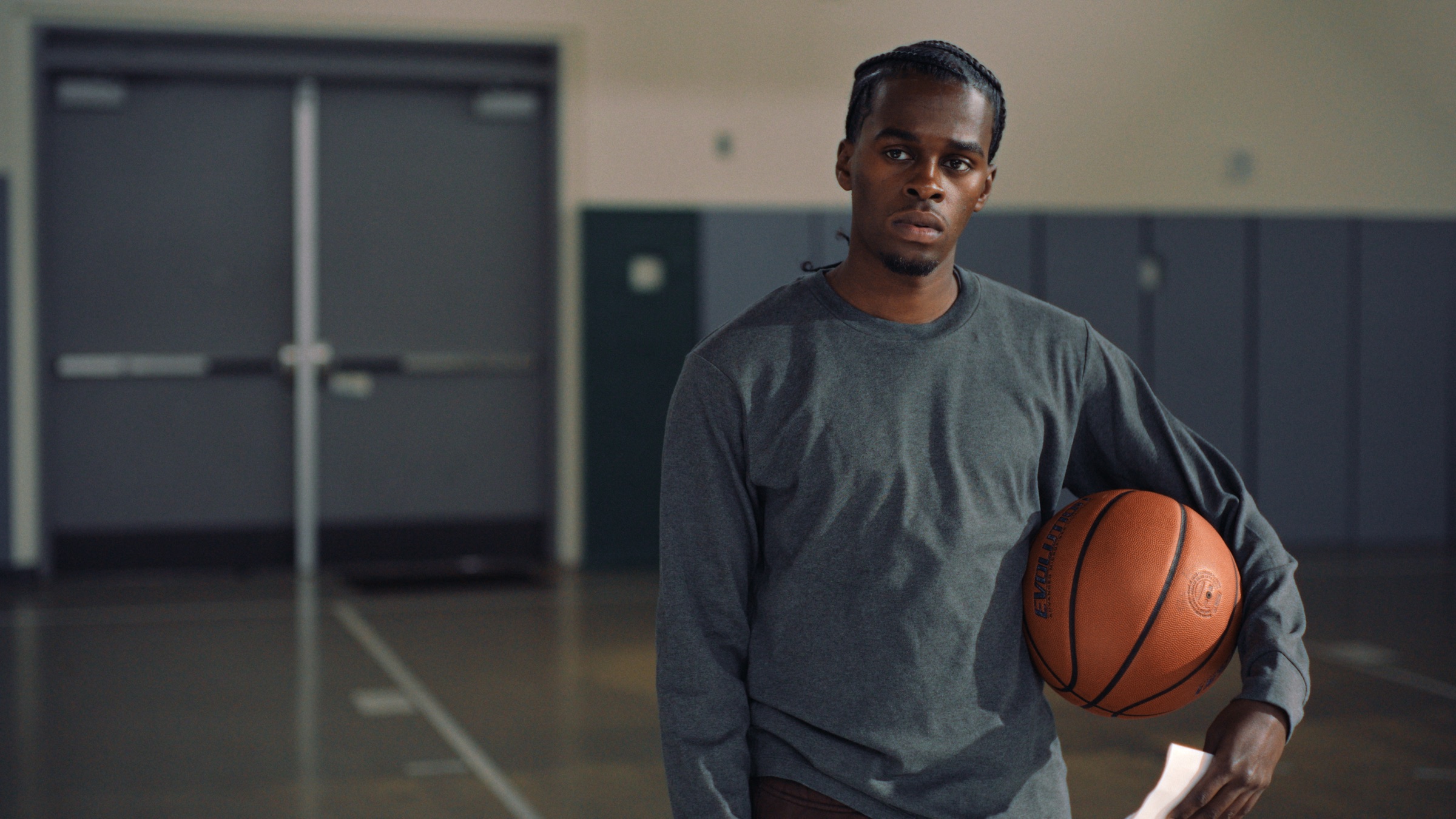 Hassan stands alone in a school gym, holding a basketball under his arm and looking straight at the camera.