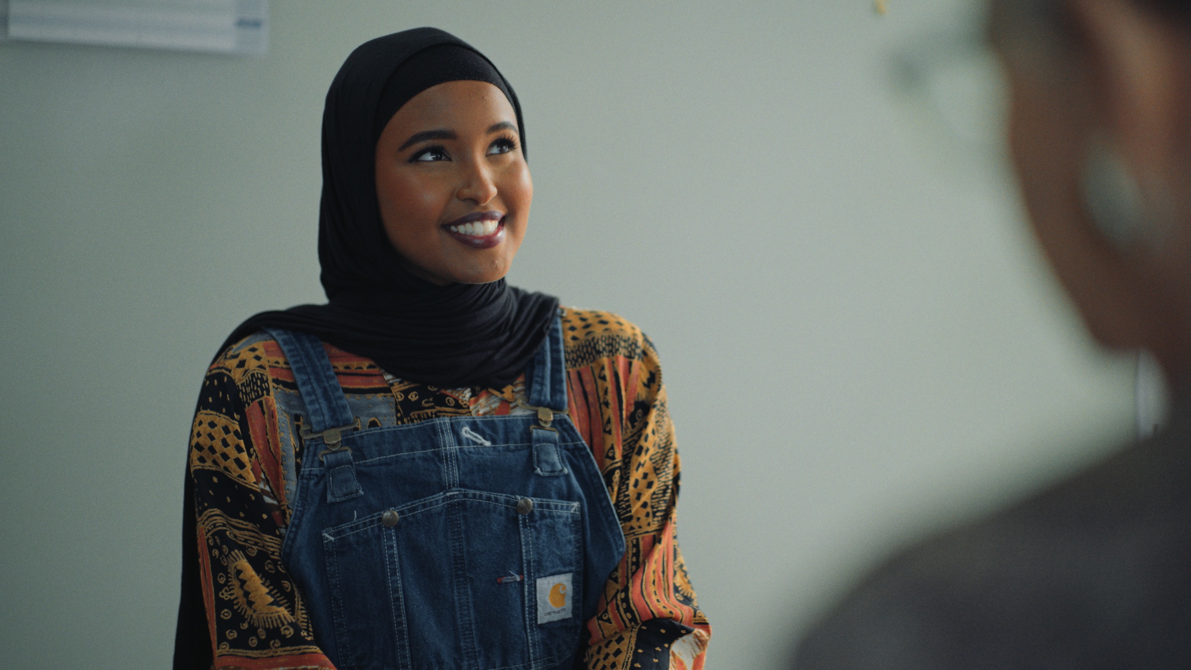 Ibtisam in denim overalls and a patterned shirt, smiling across a desk.