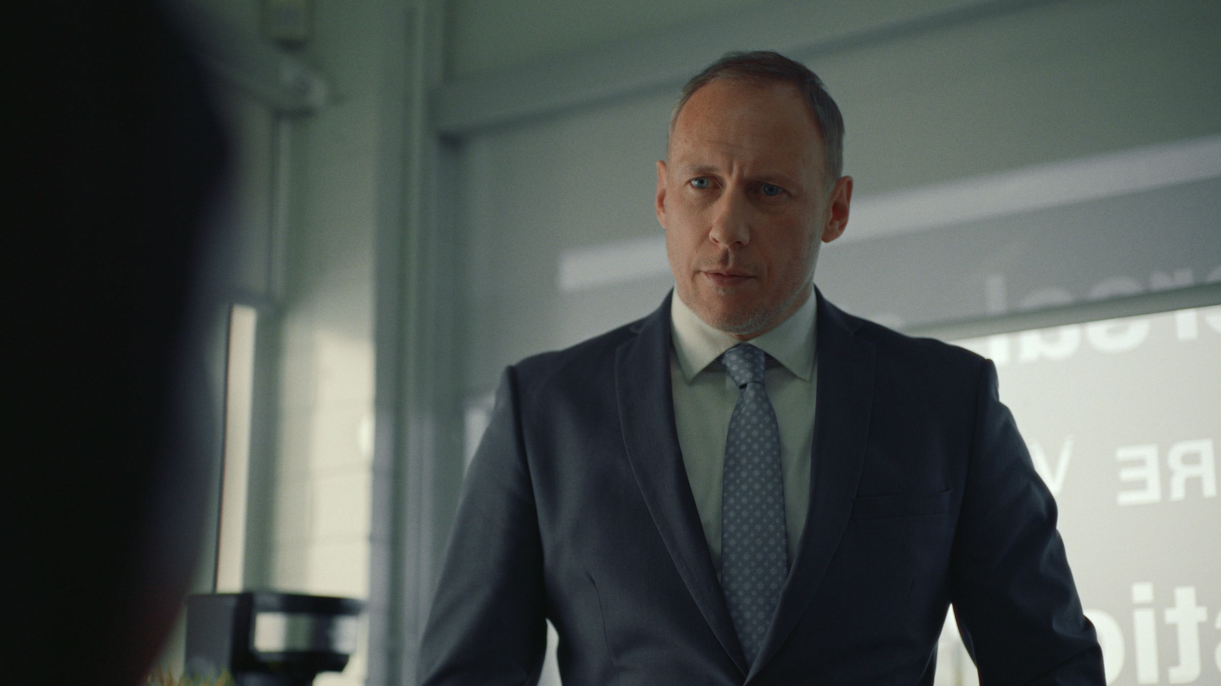 A man in a navy suit and tie, standing in an office.